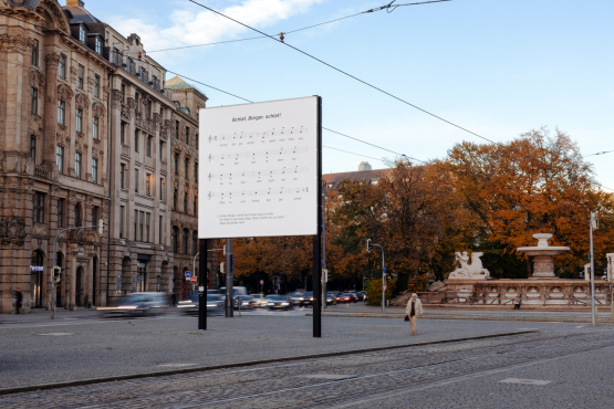 Diagonal view of the billboard at Lenbachplatz. The musical notation of the nursery rhyme "Schlaf, Kindlein, schlaf!" (Sleep, my child, sleep") appears in black lettering on a white background with the lyrics changed to "Schlaf, Bürger, schlaf!" ("Sleep, my fellow German, sleep!").